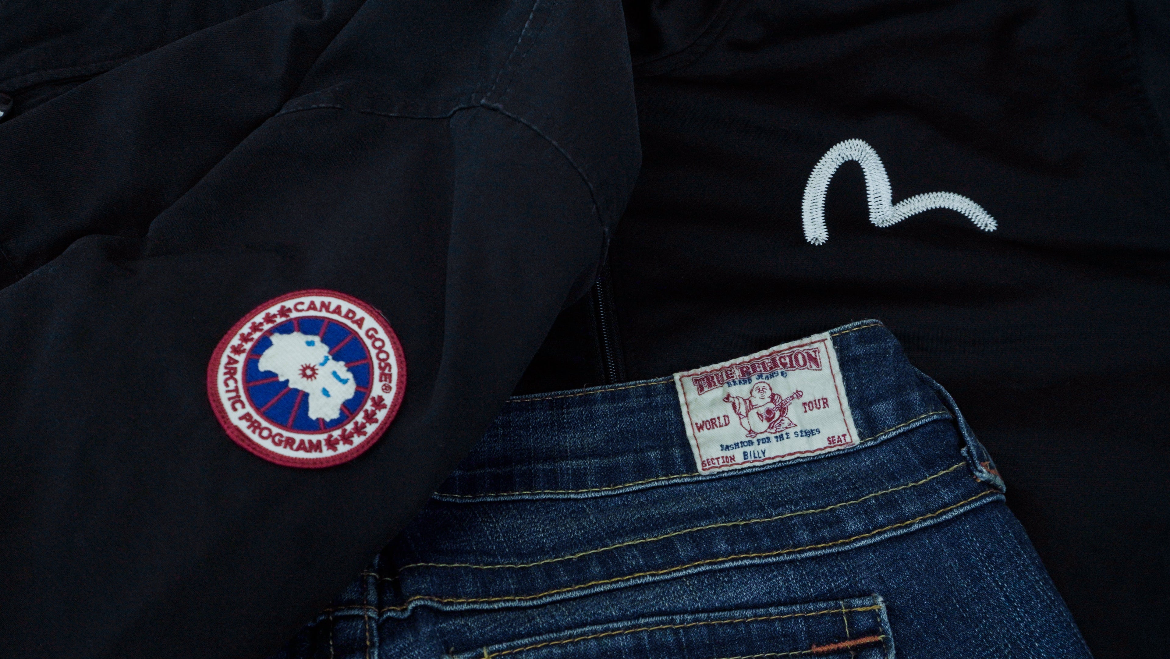 Black jacket with a Canada Goose logo, black jacket with an Evisu logo and blue jeans with a True Religion label